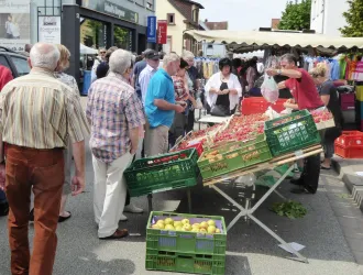 Ein Marktplatz mit vielen Menschen, die frische Produkte kaufen. Obst und Gemüse sind in Kisten arrangiert, während Verkäufer mit den Kunden interagieren.