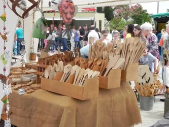 Ein Marktstand mit vielen handgemachten Holzutensilien steht im Vordergrund. Im Hintergrund sind viele Besucher zu sehen, die sich umsehen.