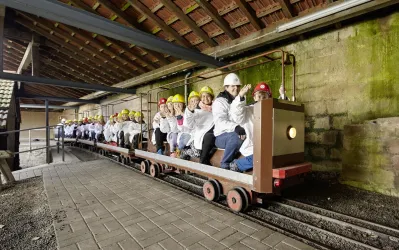 Eine Gruppe von Menschen fährt auf einer kleinen Bahn in einem unterirdischen Gang. Sie tragen Helme und lachen, während sie auf der Fahrt sitzen.