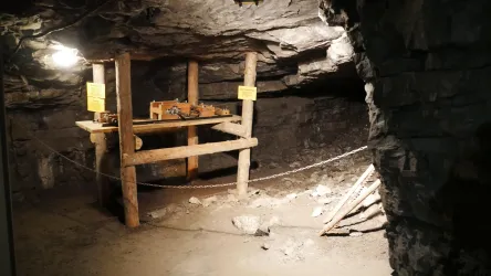 Ein unterirdischer Raum mit einem Holzgestell, auf dem Modelle ausgestellt sind. Die Wände bestehen aus grauem Gestein und es gibt schwaches Licht.