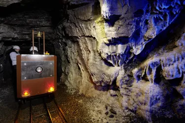 Ein Schacht mit einer kleinen Lokomotive auf Schienen und zwei Arbeitern. Die Wände sind mit interessanten mineralischen Formationen und blauer Beleuchtung dekoriert.
