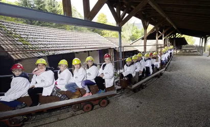 Eine Gruppe von Kindern sitzt in einer Bahn unter einem Holzgebäude. Sie tragen Sicherheitshüte und weiße Mäntel.