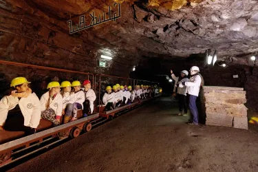 Eine Gruppe von Menschen in Sicherheitshelmen sitzt auf einer Schiene im Bergwerk. Ein Führer erklärt etwas und zeigt auf die Umgebung.