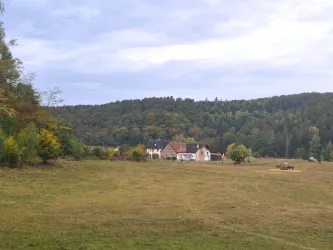 Eine ruhige Landschaft mit einem weiten Feld und sanften Hügeln im Hintergrund. Im Vordergrund sind einige Gebäude und Bäume sichtbar.