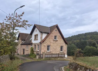 Ein charmantes Haus aus Stein in einer ländlichen Umgebung. Im Hintergrund sind Bäume und eine sanfte Hügelkette zu sehen.