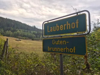 Ein Wegweiser zeigt die Namen "Lauberhof" und "Gutenbrunnerhof". Im Hintergrund ist eine grüne Wiese mit Bäumen zu sehen.