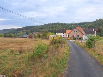 Eine ländliche Straße führt zu einem kleinen Dorf mit traditionellen Häusern. Im Hintergrund sind sanfte Hügel und ein bewölkter Himmel zu sehen.