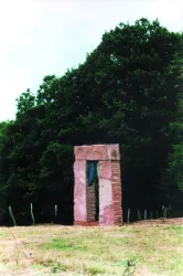 Ein steinerner Torbogen steht in einer Wiese, umgeben von Bäumen. Das Kunstwerk zeigt eine interessante Kombination aus Natur und Architektur.
