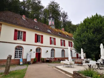 Ein historisches Gebäude mit roten Fensterläden und einem Turm. Der Gartenbereich ist mit Tischen und Stühlen ausgestattet, umgeben von Bäumen.