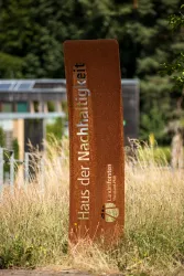 Ein rostiger Schild mit der Aufschrift „Haus der Nachhaltigkeit“ steht in einem grünen Umfeld. Im Hintergrund sind moderne Gebäude und Pflanzen sichtbar.