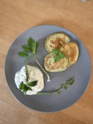 Ein Teller mit drei goldbraunen Pfannkuchen und einer Beilage aus Joghurt. Frische Kräuter dekorieren das Gericht und verleihen ihm eine ansprechende Note.