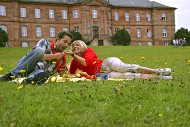 Ein Paar genießt ein Picknick auf einer Wiese, umgeben von Blumen. Im Hintergrund ist ein historisches Gebäude zu sehen.