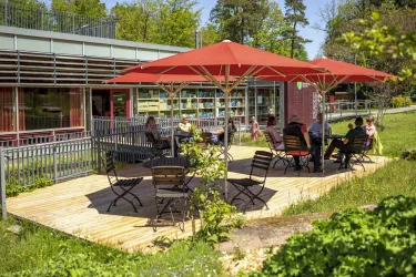 Eine gemütliche Terrasse mit roten Sonnenschirmen und Holzstühlen. Menschen sitzen entspannt und genießen die sonnige Umgebung.