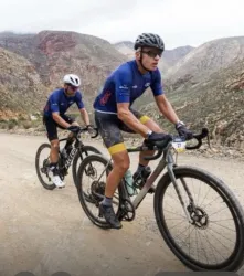 Zwei Radfahrer fahren auf einem schottrigen Weg durch eine gebirgige Landschaft. Die Umgebung ist rau und beeindruckend, mit hohen Bergen im Hintergrund.