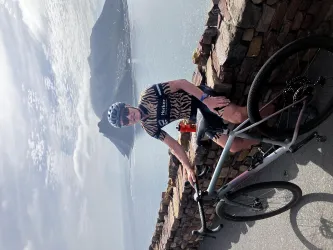 Ein Radfahrer posiert neben seinem Fahrrad mit Blick auf eine Küstenlandschaft. Der Himmel ist klar und die Umgebung wirkt malerisch.