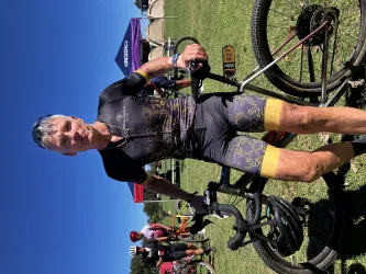 Ein Radfahrer steht neben seinem Fahrrad auf einer Wiese. Im Hintergrund sind weitere Teilnehmer und ein blauer Himmel zu sehen.