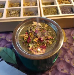 Eine Teekanne mit einer Mischung aus verschiedenen Kräutern und Blüten auf einem feinen Sieb. Im Hintergrund sind weitere Kräuter in kleinen Boxen sichtbar.