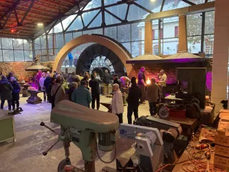Eine Gruppe von Menschen steht in einer industriellen Umgebung mit Werkstattausrüstung. Im Hintergrund sind große Bögen und Lichtinstallationen zu sehen.