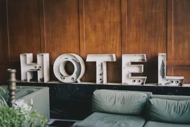 Das Bild zeigt das Wort "HOTEL" aus großen, rustikalen Buchstaben vor einer Holzwand. Im Vordergrund sind bequeme grünen Sofas zu sehen.