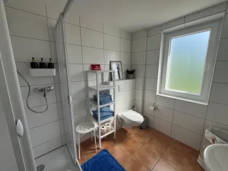 Ein modernes Badezimmer mit weißen Fliesen und einer Dusche. Auf einem Regal stehen Handtücher und Pflegeprodukte.
