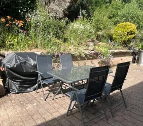 Ein gemütlicher Gartentisch mit Stühlen steht auf einer gepflasterten Fläche. Im Hintergrund sind bunte Blumen und grünes Gestrüpp zu sehen.
