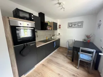 Eine moderne Küche mit schwarzen Schränken und Holzdetails. Der Raum ist hell und verfügt über einen kleinen Esstisch und Stühle.