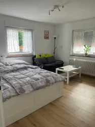 Ein gemütliches Zimmer mit einem Bett, einer schwarzen Couch und einem Tisch. Große Fenster lassen viel Tageslicht herein.