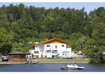 Ein ruhiger See mit einem Boot und einer weißen Unterkunft am Ufer. Dahinter sind Bäume und eine grüne Landschaft zu sehen.