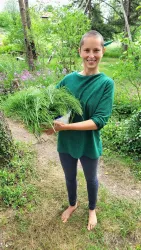 Eine Person steht barfuß im Freien und hält frische Kräuter in der Hand. Im Hintergrund sind grüne Pflanzen und Blumen zu sehen.