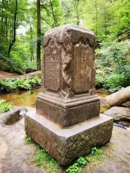 Ein alter Steinsockel mit eingravierten Inschriften steht in einem grünen Wald. Im Hintergrund fließt ein kleiner Bach.