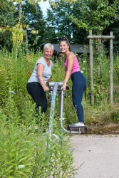 Zwei Frauen trainieren zusammen an einem Fitnessgerät im Freien. Sie lächeln und haben Spaß in einer grünen Umgebung.