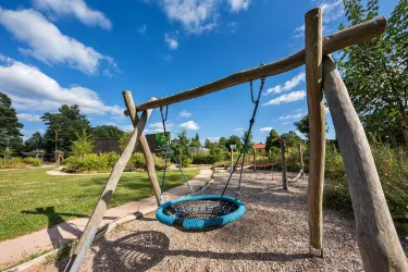 Eine große Nestschaukel aus Holz steht auf einem Spielplatz. Der Himmel ist blau und es gibt grüne Bäume im Hintergrund.