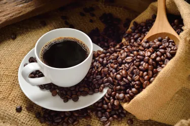 Eine Tasse frischer Kaffee steht neben einer Tüte voller Kaffeebohnen. Die Bohnen sind teils ausgekippt und liegen auf einem rustikalen Untergrund.