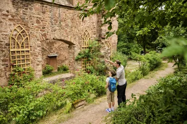 Ein Erwachsener zeigt einem Kind eine alte, verfallene Mauer mit Fenstern. Umgeben von einer grünen Gartenlandschaft wirkt die Szene ruhig und einladend.