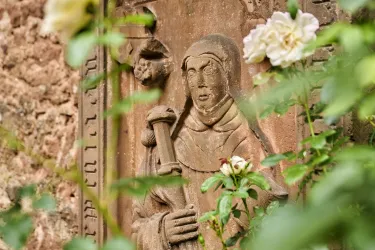 Eine Steinreliefdarstellung einer Nonne, umgeben von grünen Pflanzen und weißen Rosen. Im Hintergrund ist eine historische Wand zu sehen.