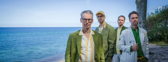 Vier Männer stehen am Strand in bunten Outfits. Im Hintergrund sieht man das klare Wasser und den blauen Himmel.