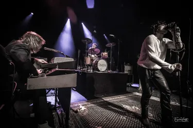 Eine Live-Band tritt auf einer dunklen Bühne auf. Der Sänger interagiert mit dem Publikum, während der Keyboarder spielt.
