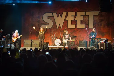 Eine Rockband tritt auf einer Bühne vor einem begeisterten Publikum auf. Im Hintergrund ist das große Logo "SWEET" zu sehen.
