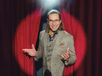 Ein Mann mit langen Haaren steht im Spotlight und lächelt. Er trägt einen eleganten Anzug und steht vor einem roten Vorhang.