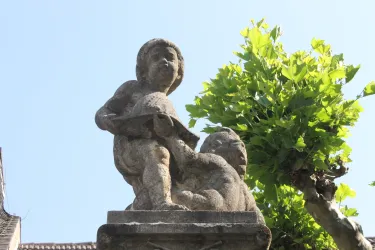 Eine Statue von zwei Kindern, die auf einem Sockel stehen. Im Hintergrund sind grüne Blätter eines Baumes sichtbar.
