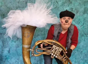 Ein clownmäßiger Künstler mit einer Tuba und einem großen, weißen Federschmuck steht vor einem bunten Hintergrund. Der Künstler trägt eine rote Bluse und sieht humorvoll aus.