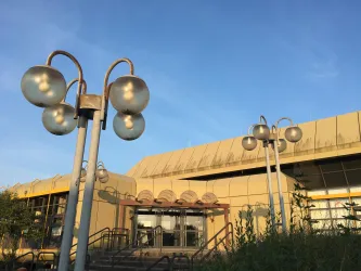 Ein modernes Gebäude mit einer gelben Fassade und großen Fenstern. Im Vordergrund befinden sich stilvolle Lampen und Grünflächen.