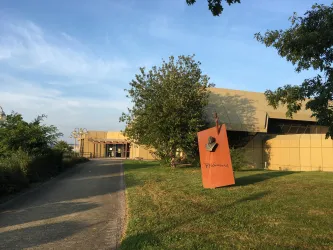 Ein modernes Gebäude mit gelber Fassade und einer großen, roten Skulptur im Vordergrund. Der Weg führt zu dem Eingang, umgeben von Bäumen und Gras.