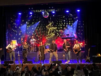 Eine lebhafte Band spielt auf einer Bühne vor einem begeisterten Publikum. Farbiges Licht und Dekorationen sorgen für eine festliche Atmosphäre.