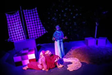 Eine Szene aus einem Theaterstück mit einer surrealen Umgebung. Drei Schauspieler in farbenfrohen Kostümen stehen und sitzen vor einem Schiff mit gestreiften Segeln.