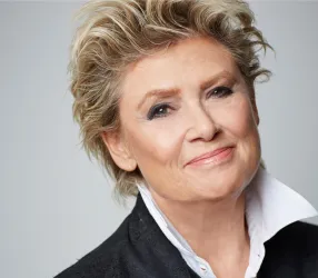 Ein lächelnde Frau mit kurzen, blonden Haaren und einem stilvollen Outfit. Sie hat einen freundlichen Gesichtsausdruck und trägt eine weiße Bluse mit einem schwarzen Blazer.
