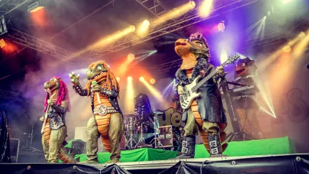Eine Band in Dinosaurier-Kostümen spielt auf einer Bühne. Bunte Lichter und Nebel schaffen eine festliche Atmosphäre.
