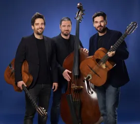 Drei Musiker stehen in eleganter Kleidung vor einem blauen Hintergrund. Einer hält eine Gitarre, ein anderer einen Kontrabass und der dritte eine Gitarre in der Hand.
