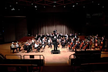 Ein Orchester spielt auf einer Bühne mit einem Dirigenten. Die Musiker sitzen in typischer Anordnung mit verschiedenen Instrumenten.