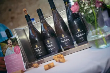 Auf dem Tisch stehen mehrere Weinflaschen mit schwarzen Etiketten. Daneben liegt eine Vase mit frischen Blumen und einige Korken.